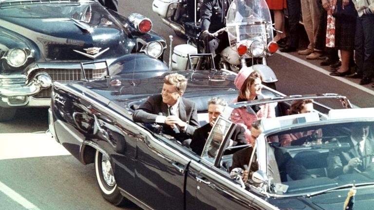 "JFK Revisited - Die Wahrheit über den Mord an John F. Kennedy": Zwei Männer und eine Frau sitzen lachend und winkend in einem dachlosen Auto während die Menge ihnen zujubelt.