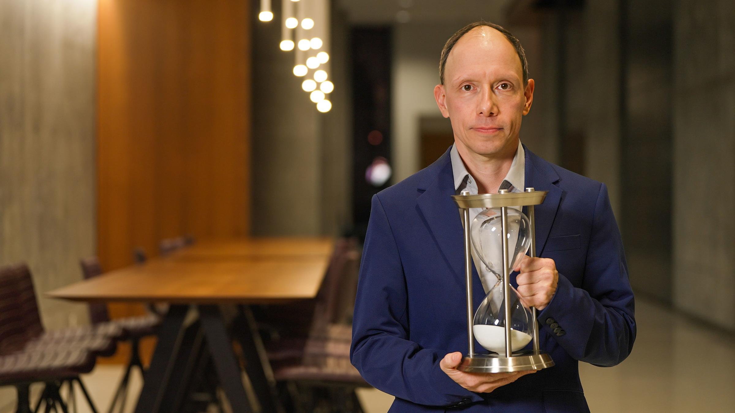 ZDF.zeit: "Forever Young - Wie können wir das Altern stoppen?": In der Bildmitte steht der deutsche Forscher Steve Horvath. Er trägt ein blaues Jackett und hält eine große Sanduhr in den Händen.