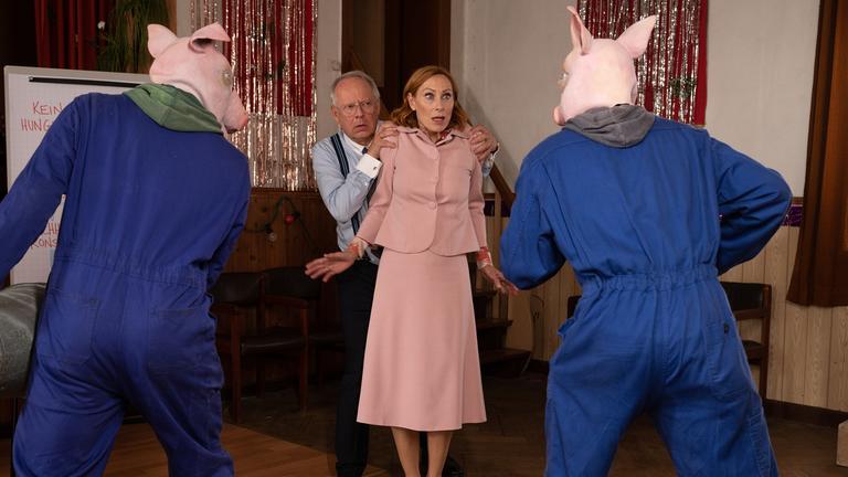 "Familie Bundschuh - Bundschuh vs. Bundschuh": Zwei bedrohlich wirkende Männer, die bösartige Schweinemasken und  Blaumänner tragen, bedrohen Gundula (Andrea Sawatzki) und Gerald Bundschuh (Axel Milberg). Das Paar steht elegant bekleidet zwischen den beiden Aktivisten in einem mit Lamettavorhängen geschmückten Saal. Gerald versteckt sich ängstlich hinter der  erschrockenen blickenden Gundula, die ihre Hände abwehrend und Gerald schützend zur Seite gestreckt hat.