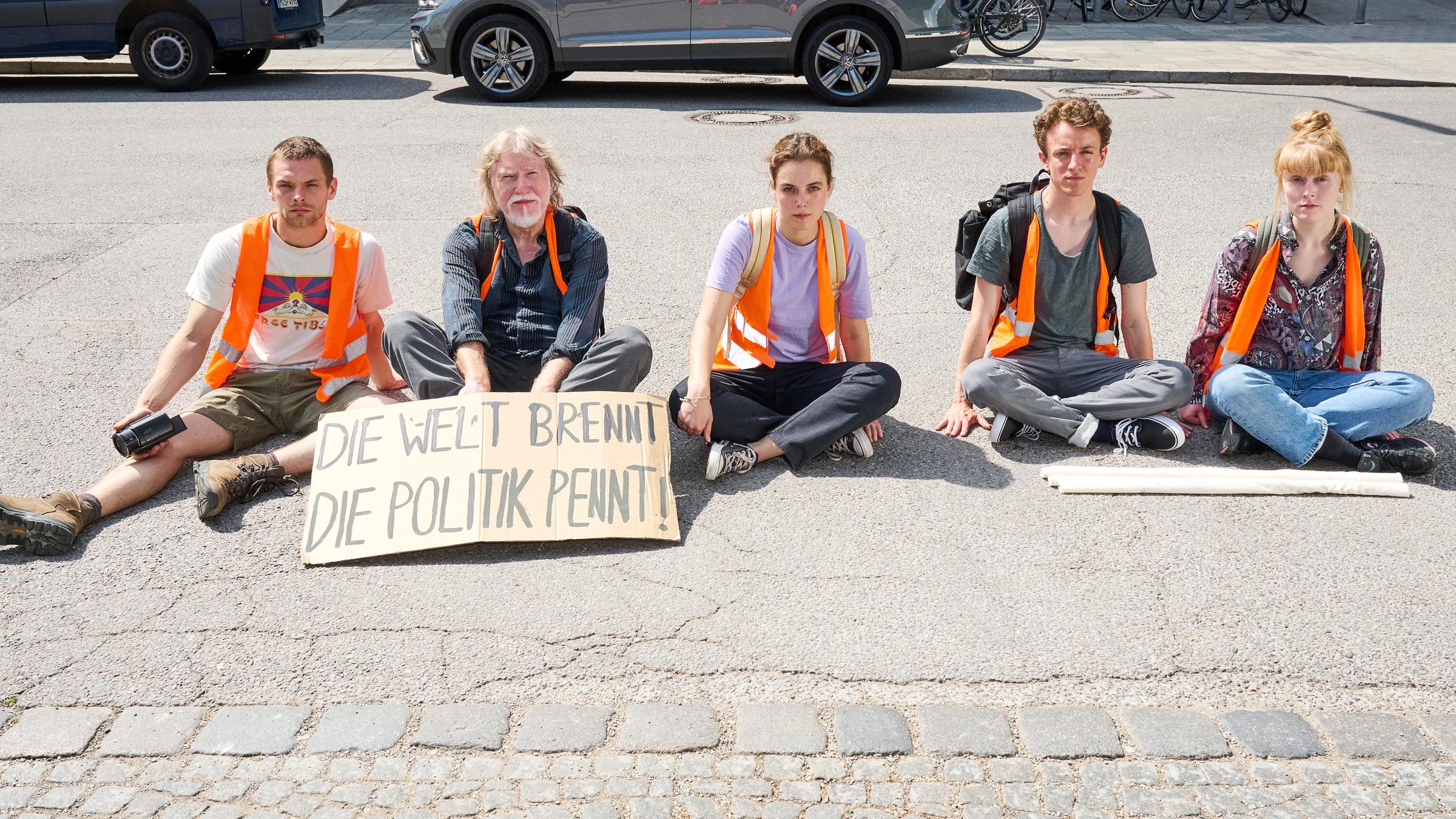 "Aufgestaut - Lew": Franzi (Maria Goletz), Finn (Adrian Grünewald), Lena (Valerie Stoll), Henning (Joachim Seemann) und Anatol (Valentino Dalle Mura) tragen Warnwesten und sitzen nebeneinander auf der Straße. Vor Henning liegt ein Schild auf dem steht: "Die Welt brennt. Die Politik pennt!"