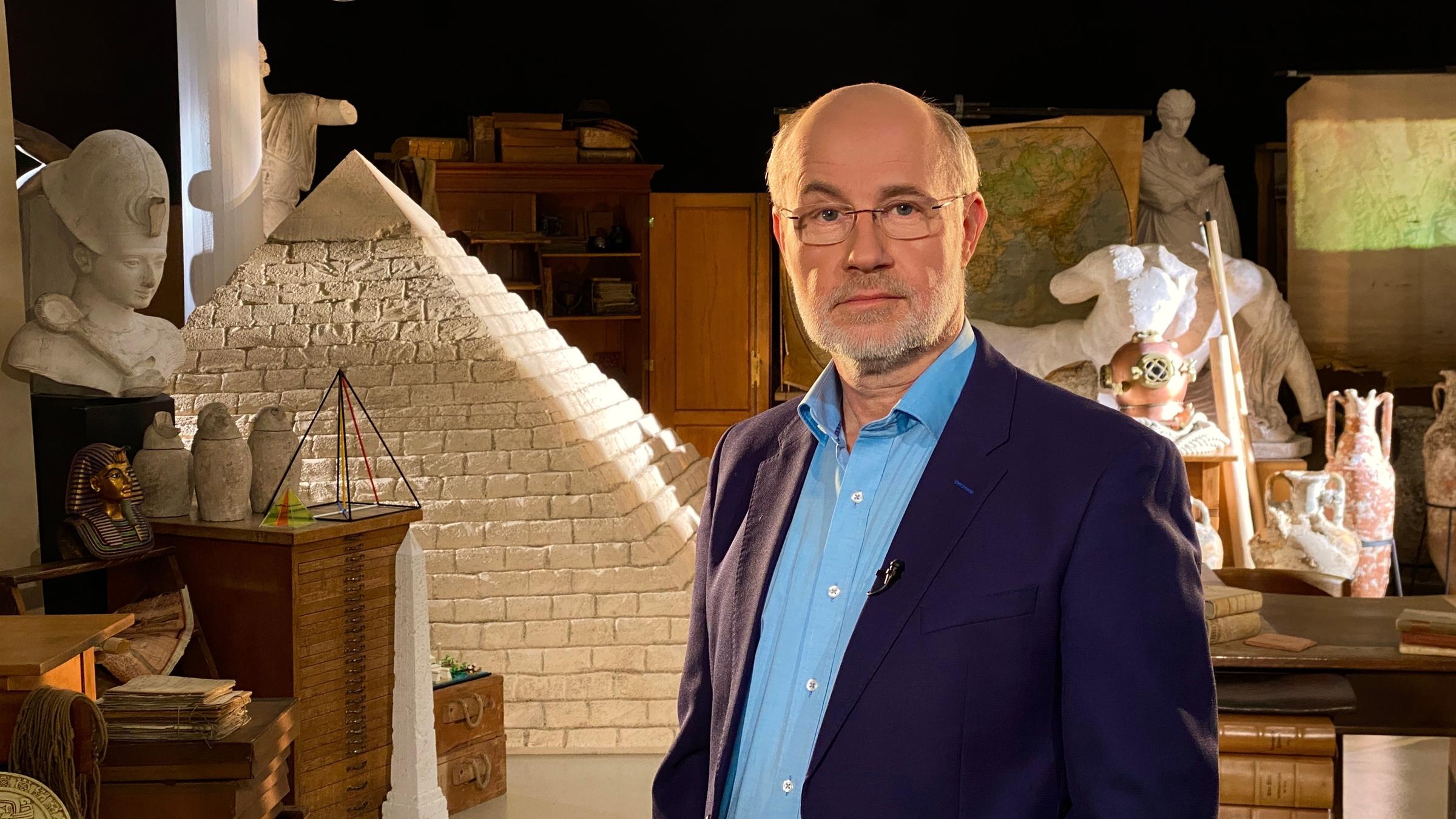 "Ungelöste Fälle der Archäologie - Pyramiden": Der Moderator Harald Lesch in einem Raum mit Nachbildungen diverser Artefakte aus der Antike, darunter ein großes Modell einer Pyramide.