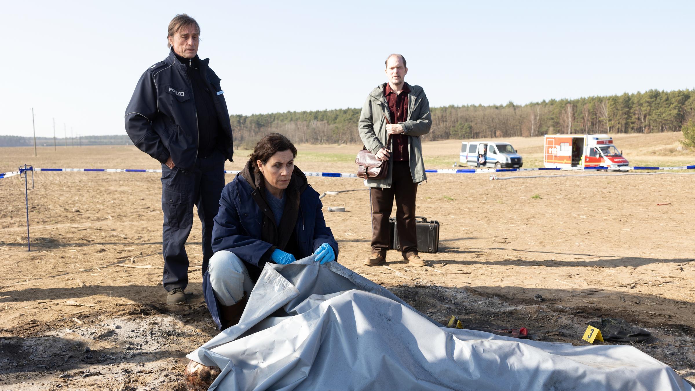 "Das Mädchen von früher": Eine abgedeckte Leiche liegt auf einem trockenen Feld. Kriminalhauptkommissarin Maria Voss (Nina Kunzendorf) kniet daneben. Ihre Kollegen Lars Klapproth (André Hennicke) und Teo Kromann (Godehard Giese) stehen hinter ihr.