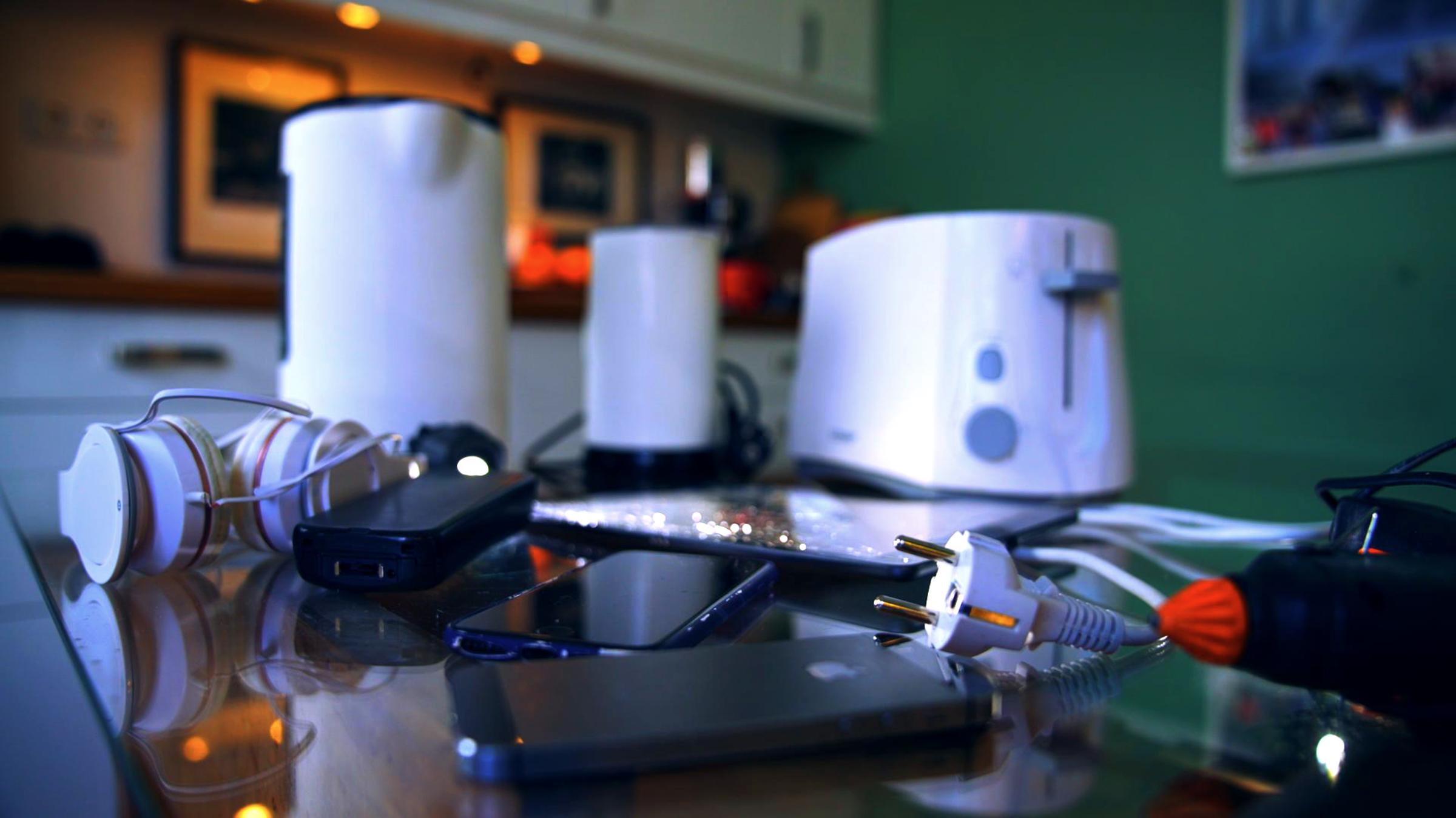 Mehrere kaputte Elektrogeräte stehen und liegen auf einem Tisch: ein Wasserkocher, ein Toaster, Kopfhörer und Smartphones.