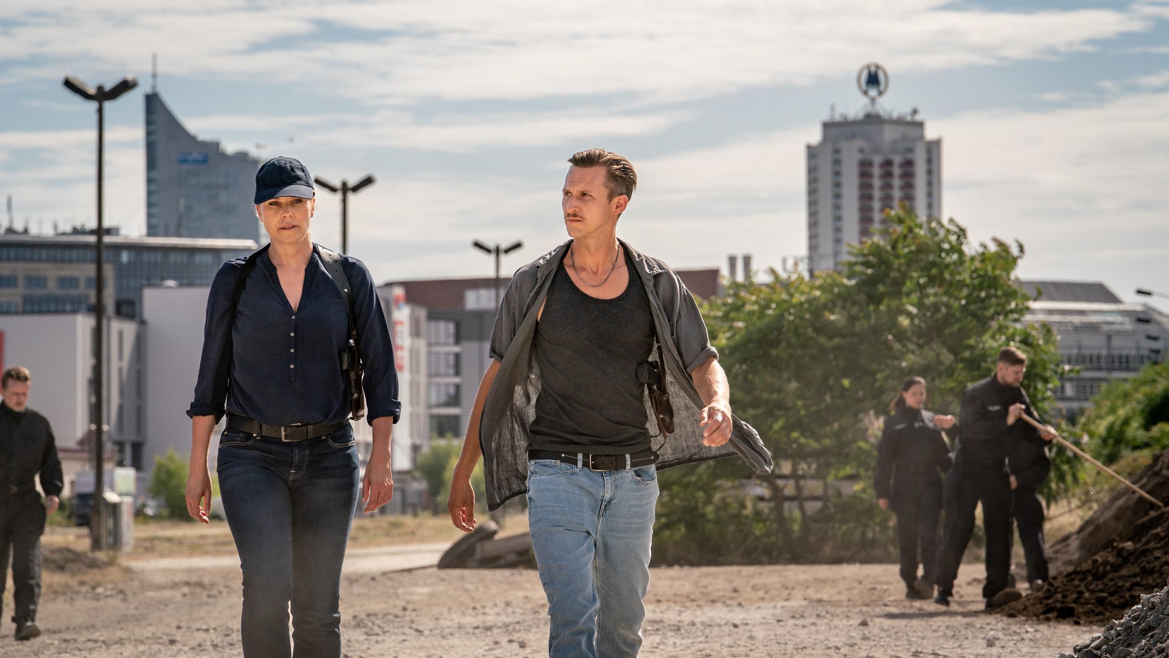 "SOKO Leipzig - Joela": Moritz Brenner (Johannes Hendrik Langer) und Ina Zimmermann (Melanie Marschke) schreiten über ein Schotterfeld, im Hintergrund stehen einige Gebäude der Stadt Leipzig. Um sie herum sind mehrere Kriminalbeamte (Komparsen) zu sehen, einer von ihnen zieht gerade ein Absperrband.