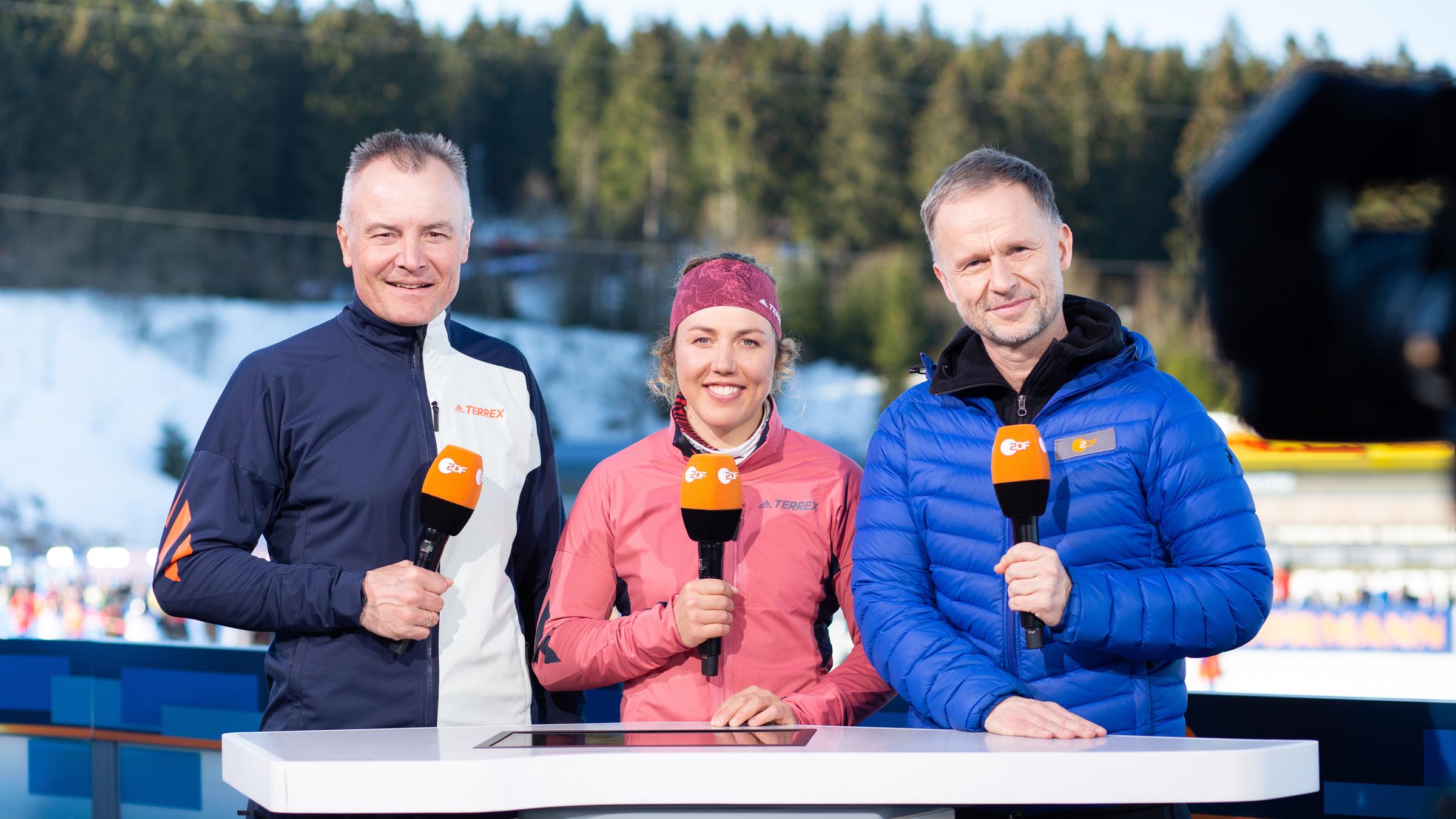 ZDF-Biathlon-Experten Sven Fischer (l.) und Laura Dahlmeier, ZDF-Moderator Alexander Ruda stehen an der Presenter-Position hinter dem Moderatiionstisch mit Mikrofonen in der Hand am Rennsteig im Oberhof.