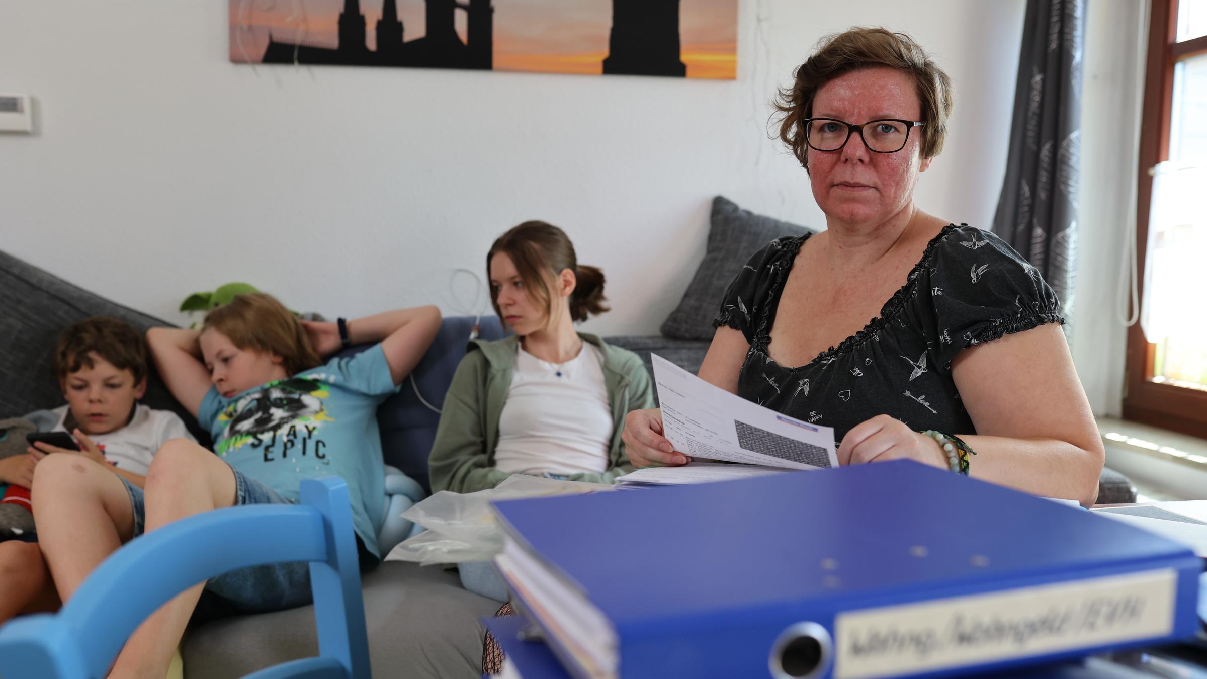 "ZDF.reportage: Armes reiches Deutschland - Alleinerziehend und abgehängt": Die Alleinerziehende Madlen Fahrenbach arbeitet am Schreibtisch, vor ihr stapeln sich Aktenordner, im Hintergrund sitzen ihre Kinder auf dem Sofa.