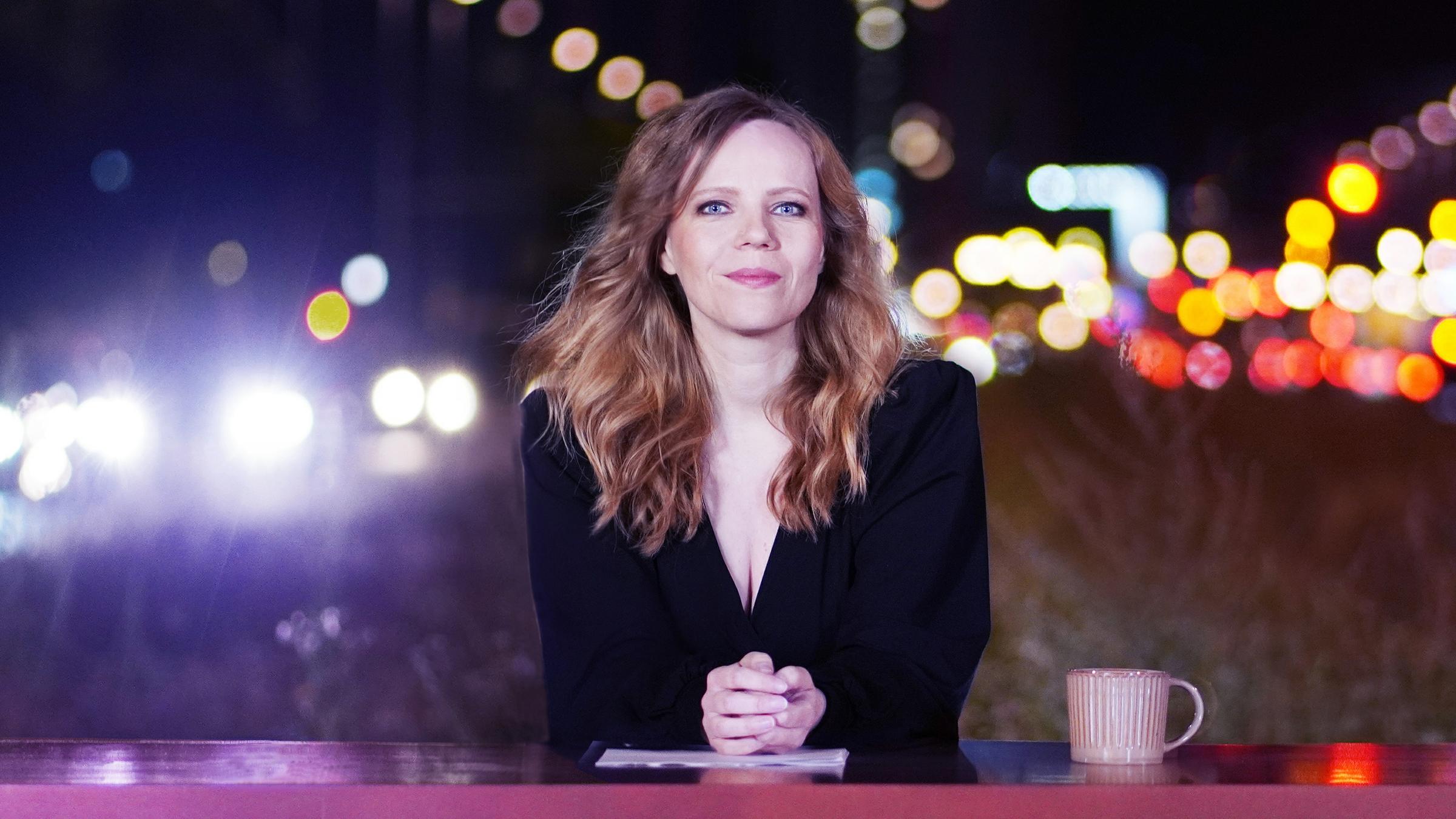 "Bosetti Late Night": Sarah Bosetti sitzt hinter einem pinkfarbenen Tresen. Sie schaut freundlich in die Kamera. Neben Ihr steht eine rosafarbene Kaffeetasse.