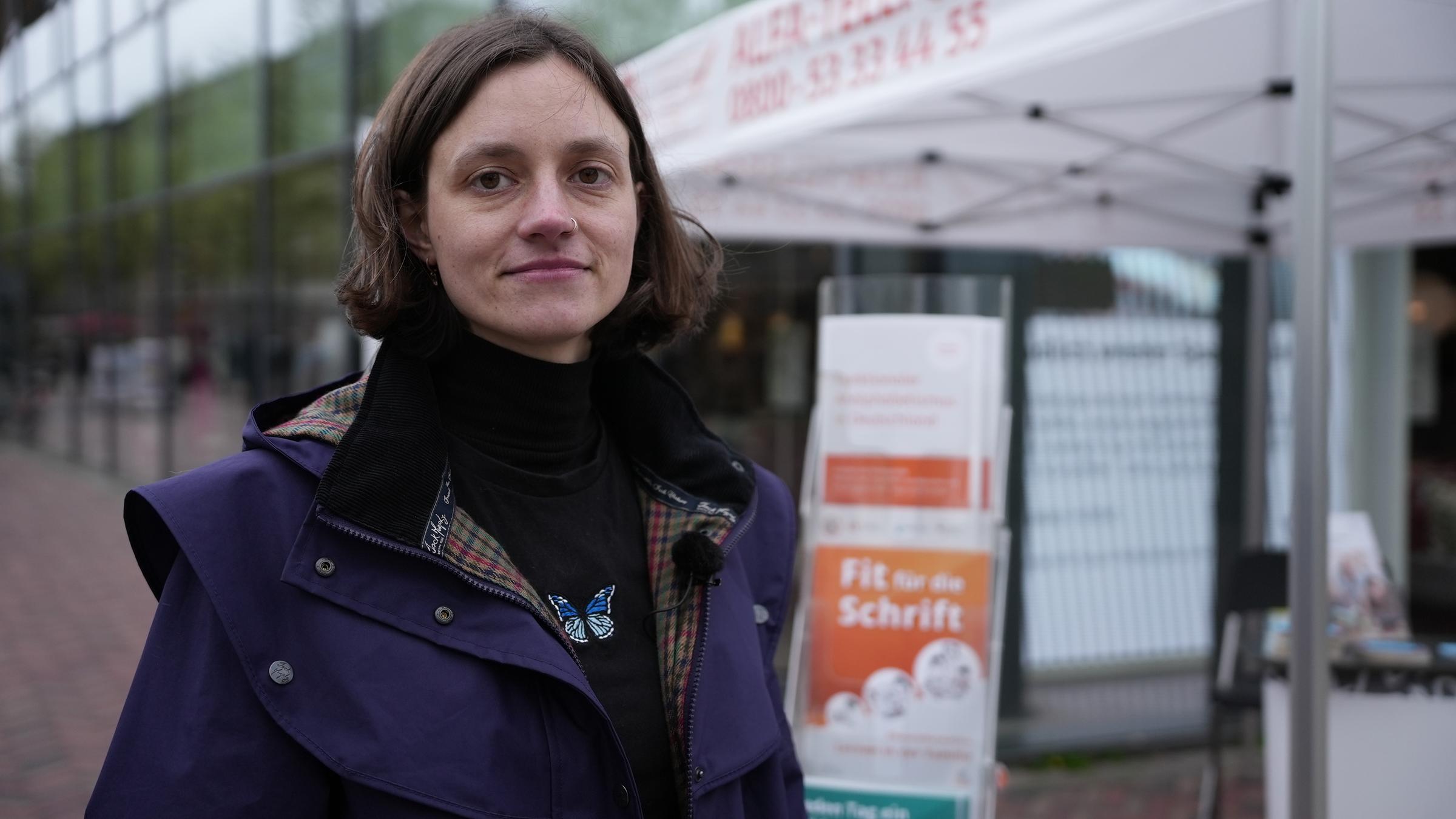 "37°: Immer Ärger mit den Buchstaben - Wenn das Alphabet zur Qual wird": Kunststudentin Lea steht vor einem Infostand, auf dem Hilfsangebote für Menschen, die nicht richtig lesen und schreiben können, beworben werden. Die Frau schaut selbstbewusst in die Kamera.