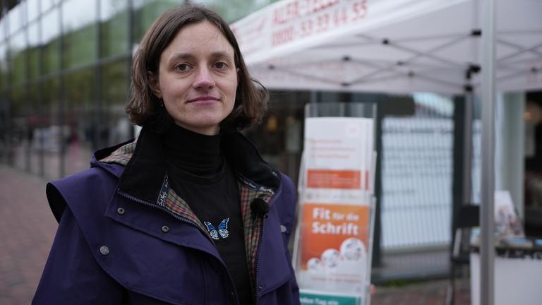"37°: Immer Ärger mit den Buchstaben - Wenn das Alphabet zur Qual wird": Kunststudentin Lea steht vor einem Infostand, auf dem Hilfsangebote für Menschen, die nicht richtig lesen und schreiben können, beworben werden. Die Frau schaut selbstbewusst in die Kamera.