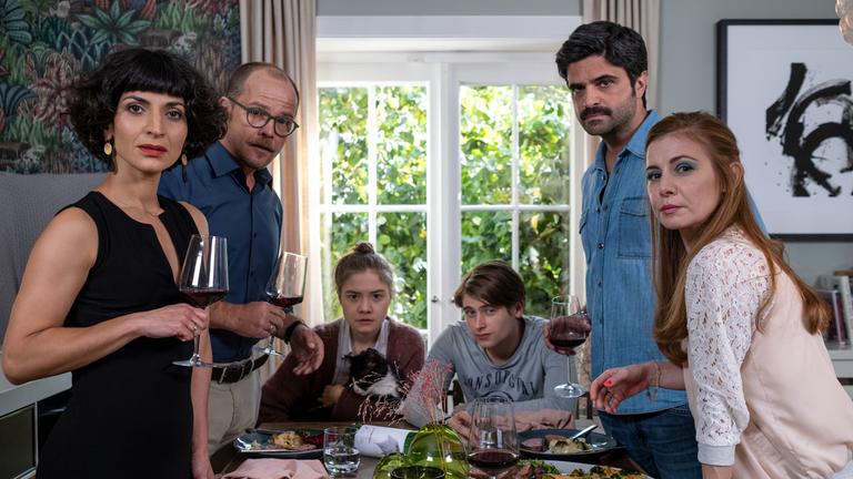 "Gäste zum Essen": Am Kopfende eines gedeckten Tisches sitzen Mila Faber (Hannah Schiller) und Leon Popov (Paul Sundheim) mit verschränkten Armen und ernstem Blick. Auf der linken Seite stehen Soraya (Neda Rahmanian) und André Faber (Matthias Koeberlin) mit Rotweingläsern in der Hand. Rechts stehen Viktor Popov (Maximilian Grill) und seine Frau Monika (Josefine Preuß). Er hält ebenfalls ein Rotweinglas in der Hand. Monika greift gerade nach einem Glas. Alle blicken ernst in die Kamera.