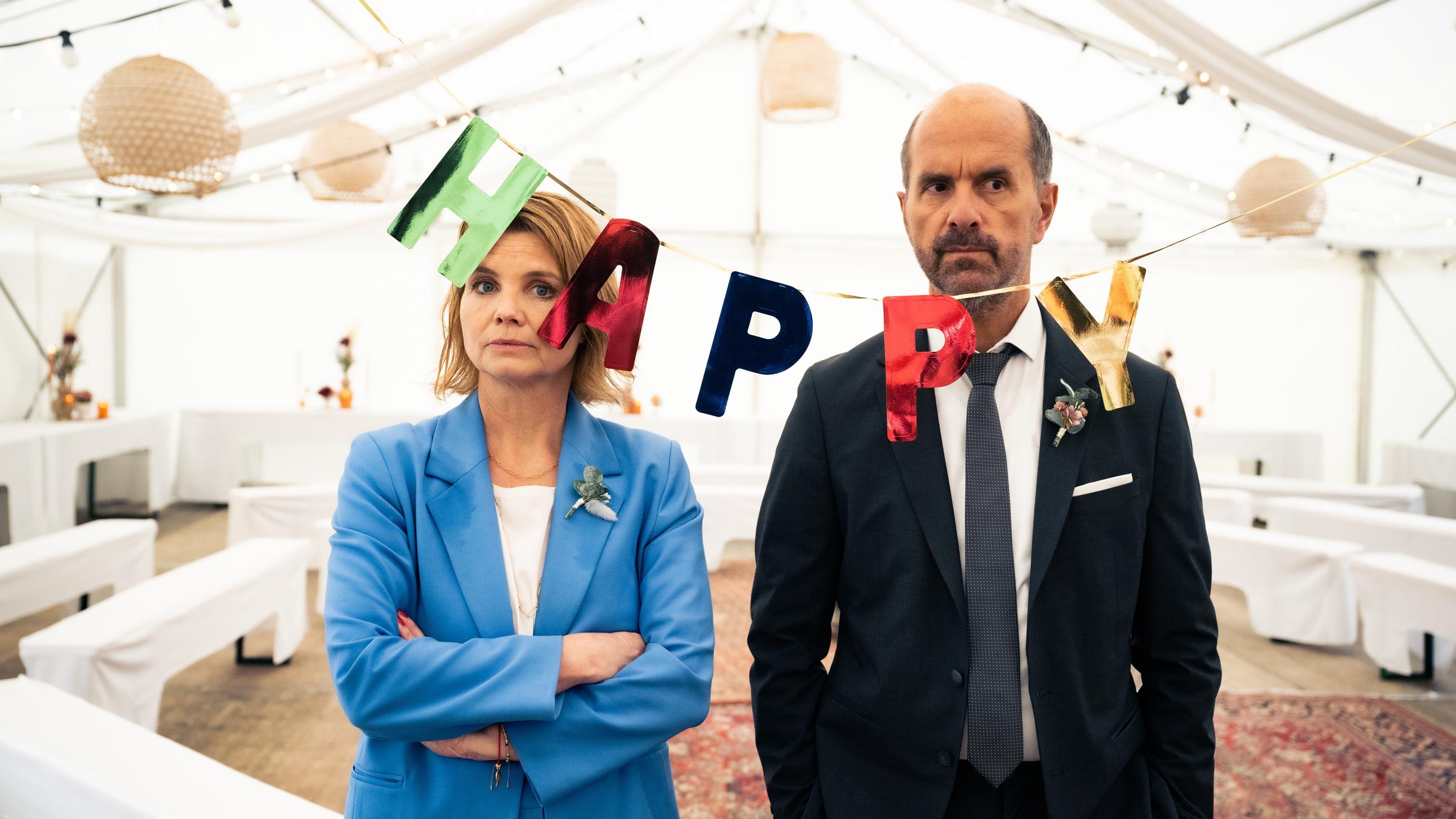 "Merz gegen Merz - Hochzeiten": Erik (Christoph Maria Herbst) und Anne (Annette Frier) stehen in Abendkleidung genervt nebeneinander. Vor ihnen hängt eine Girlande, auf der "Happy" steht und im Hintergrund ist ein weißes Hochzeitszelt mit Tischen und Bänke aufgebaut.