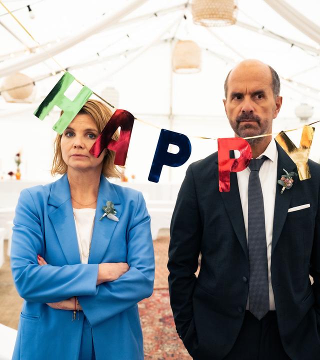 "Merz gegen Merz - Hochzeiten": Erik (Christoph Maria Herbst) und Anne (Annette Frier) stehen in Abendkleidung genervt nebeneinander. Vor ihnen hängt eine Girlande, auf der "Happy" steht und im Hintergrund ist ein weißes Hochzeitszelt mit Tischen und Bänke aufgebaut.