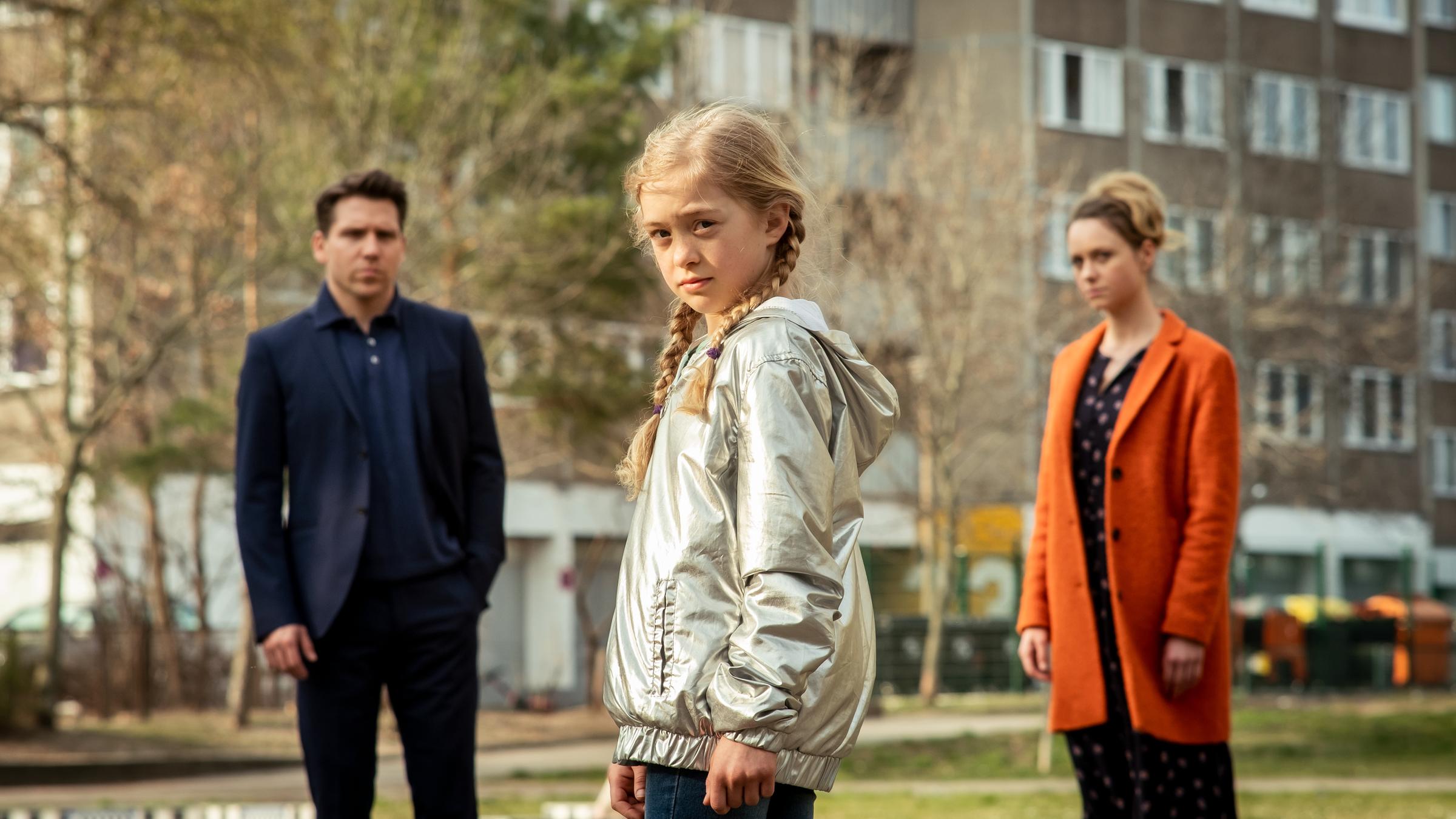 "Das bleibt unter uns": Anna (Anna Cheban), ein kleines, blondes Mädchen, blickt im Vordergrund des Bildes direkt in die Kamera, links von ihr im Bildhintergrund Alexander (Hanno Koffler) im Jacket, rechts Jana (Anna Unterberger), eine blonde Frau im orangen Mantel.