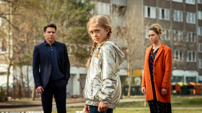 "Das bleibt unter uns": Anna (Anna Cheban), ein kleines, blondes Mädchen, blickt im Vordergrund des Bildes direkt in die Kamera, links von ihr im Bildhintergrund Alexander (Hanno Koffler) im Jacket, rechts Jana (Anna Unterberger), eine blonde Frau im orangen Mantel.