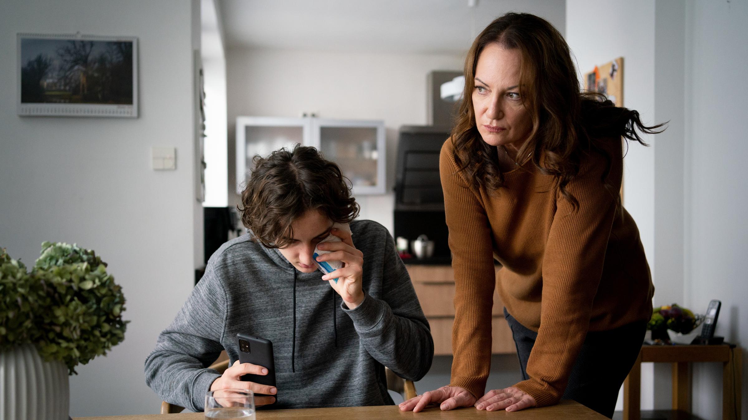 "Unter anderen Umständen - Mütter und Söhne": Jana Winter (Natalia Wörner) steht neben ihrem Sohn Leo Winter (Jacob Lee Seeliger), der mit dem Handy in der Hand an einem Tisch sitzt.