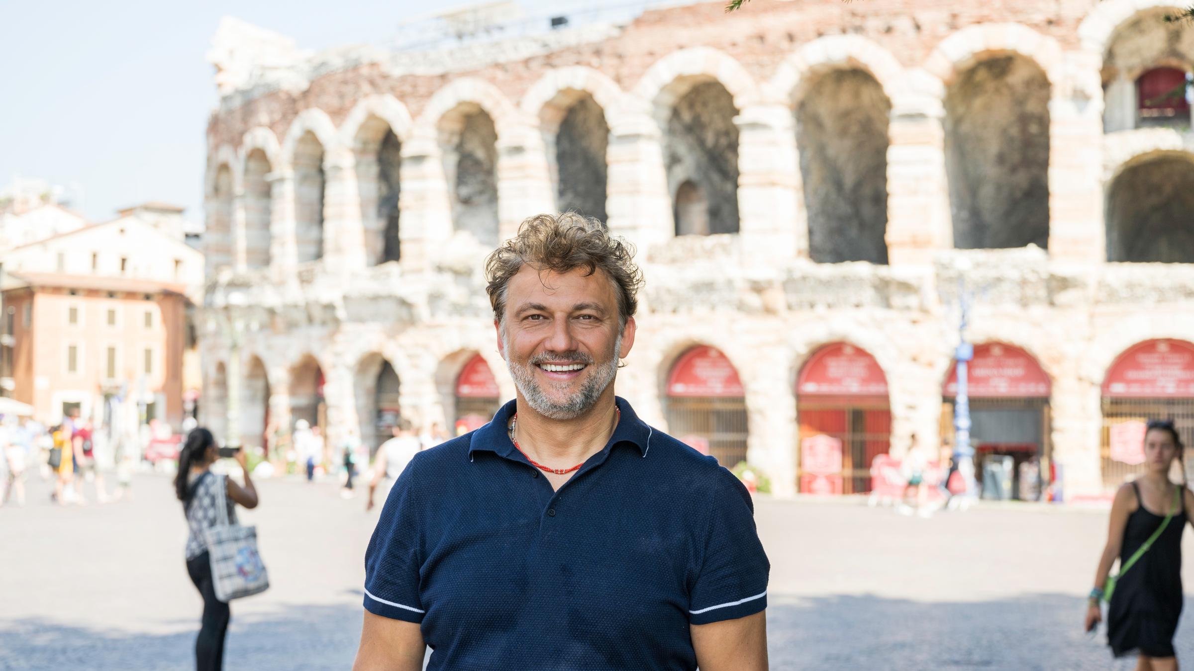 "Jonas Kaufmann in der Arena von Verona": Jonas Kaufmann steht bei sonnigem Wetter vor der Arena von Verona und lacht in die Kamera.