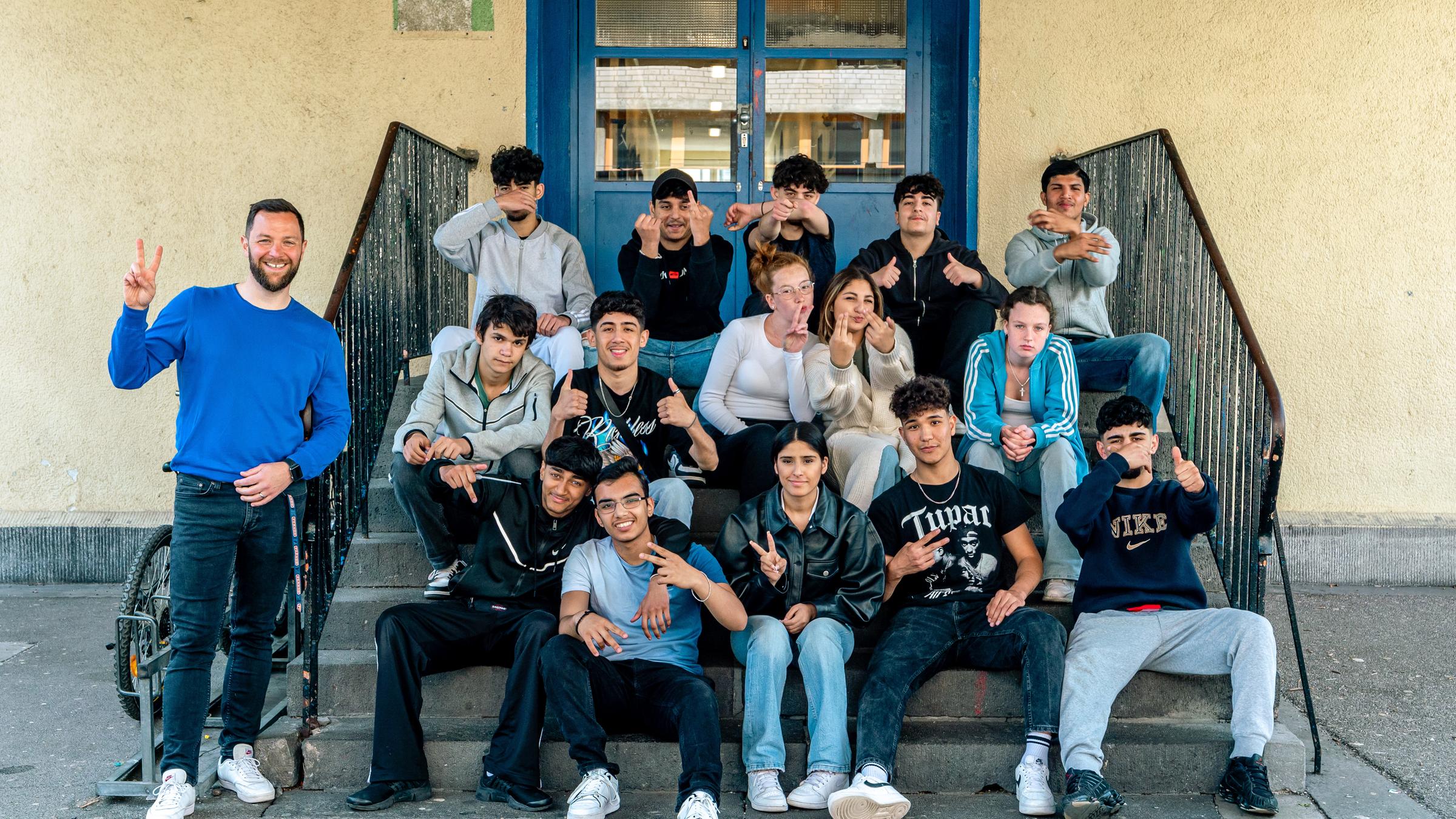 "ZDF.reportage: Hauptschulklasse 9a - Jogginghose war gestern!": Gruppenfoto: Die Schülerinnen und Schüler der Klasse 9a sitzen auf einer Treppe. An der linken Seite steht ihr Klassenlehrer. Alle machen Faxen.