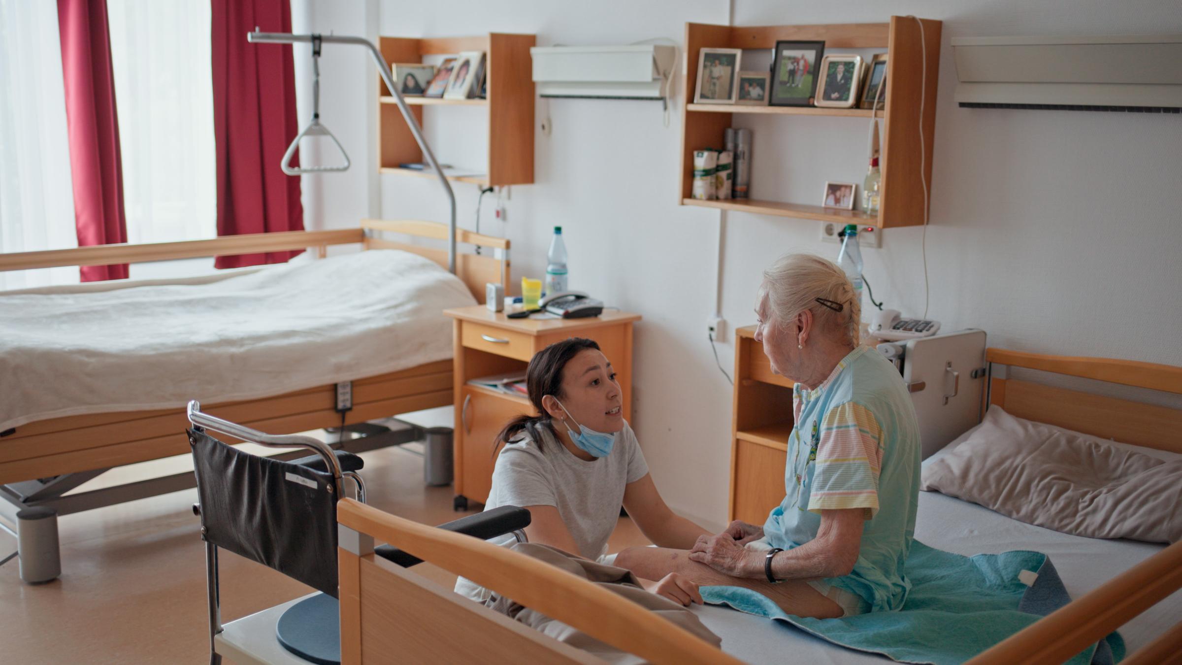 "IchDuWir - Wer pflegt wen?": Eine junge Frau in der Arbeitskleidung von medizinischem Personal sitzt vor dem Bett einer alten Frau in der Hocke und spricht mit ihr, die in einem Nachthemd aufrecht auf der Bettkante sitzt.