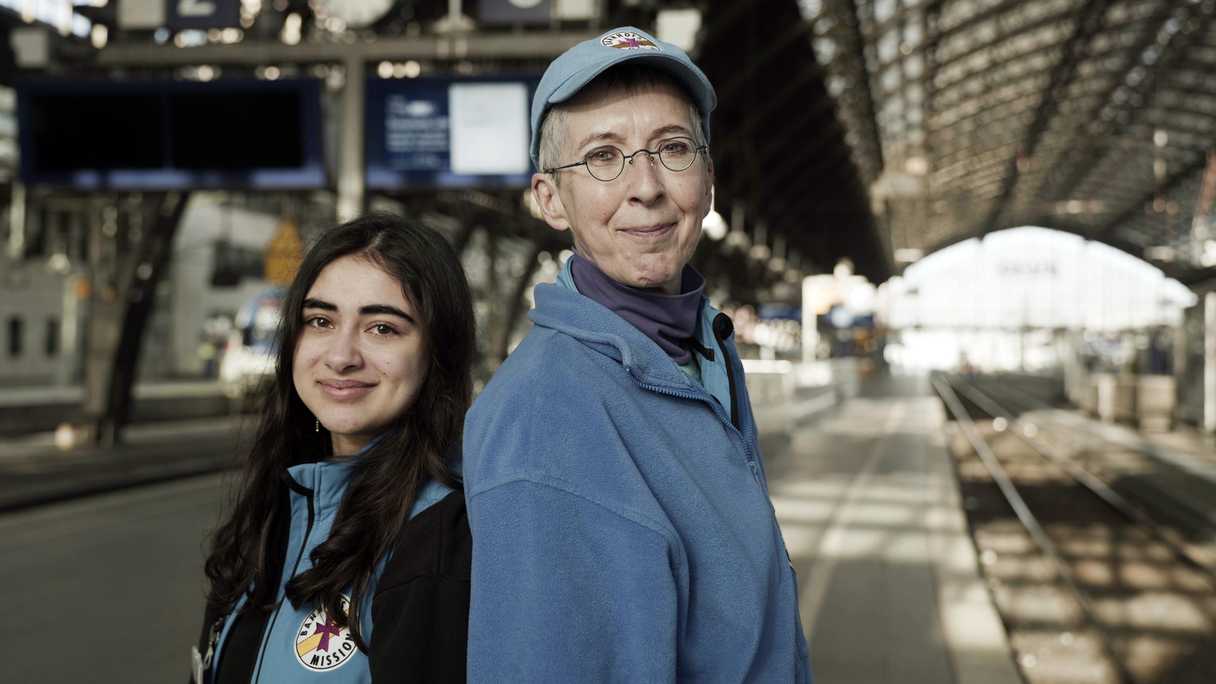 "IHR & WIR - Generationen im Konflikt": Zwei Frauen, Naina und Barbara, stehen im Kölner Hauptbahnhof Rücken an Rücken und schauen freundlich in die Kamera. Naina ist 18 Jahre alt und hat langes schwarzes Haar. Barbara ist Anfang sechzig und hat kurze, graue Haare. Sie tragen die blaue Arbeitskleidung der Bahnhofsmission.