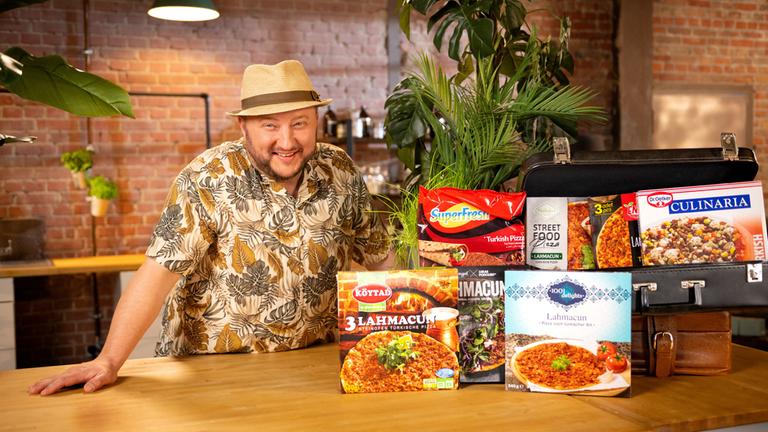 "besseresser: Urlaubsflair aus dem Supermarkt - Sebastian Lege packt aus": Sebastian Lege steht, bekleidet mit einem kurzärmligen Hemd und Strohhut, hinter der Küchentheke und lächelt in die Kamera. Vor ihm stehen diverse Packungen mit Lahmacun, einer türkischen Pizza, aus dem Supermarkt rund um zwei Koffer drapiert.