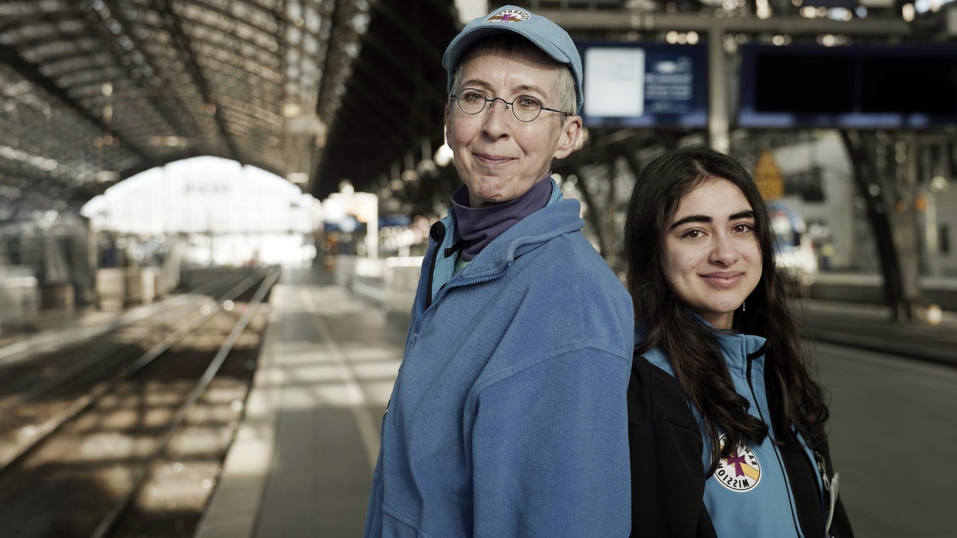 "IHR & WIR - Generationen im Konflikt": Zwei Frauen, Naina und Barbara, stehen im Kölner Hauptbahnhof Rücken an Rücken und schauen freundlich in die Kamera. Naina ist 18 Jahre alt und hat langes schwarzes Haar. Barbara ist Anfang sechzig und hat kurze, graue Haare. Sie tragen die blaue Arbeitskleidung der Bahnhofsmission.