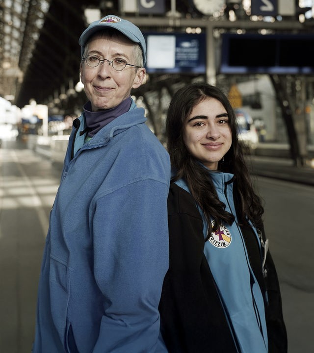 "IHR & WIR - Generationen im Konflikt": Zwei Frauen, Naina und Barbara, stehen im Kölner Hauptbahnhof Rücken an Rücken und schauen freundlich in die Kamera. Naina ist 18 Jahre alt und hat langes schwarzes Haar. Barbara ist Anfang sechzig und hat kurze, graue Haare. Sie tragen die blaue Arbeitskleidung der Bahnhofsmission.