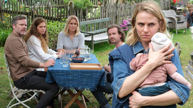 "Lena Lorenz - Vertauscht": Im Garten des Lorenzhofes sitzen Gregor (Nicholas Reinke) und Mareike Völler (Anja Antonowicz),  Lena Lorenz (Judith Hoersch) und Raffael Vollmer (Max Rothbart). Alle schauen Annika (Teresa Rizos) hinterher, die ihr Baby an sich drückt und mit besorgtem Gesichtsausdruck den Tisch verlässt.