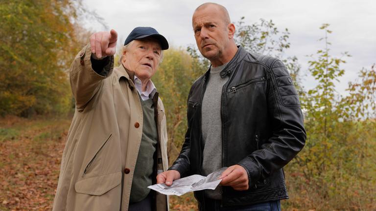 "Briefe aus dem Jenseits?": Gerd Dennert (Manfred Zapatka) und Ingo Thiel (Heino Ferch) stehen in der Natur. Ingo hat ein Bild in der Hand und und schaut aufmerksam in die Richtung, in die Gerd zeigt, während er ihm etwas zu erklären scheint.