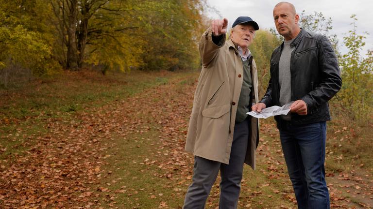 "Briefe aus dem Jenseits?": Gerd Dennert (Manfred Zapatka) und Ingo Thiel (Heino Ferch) stehen in der Natur. Ingo hat ein Bild in der Hand und und schaut aufmerksam in die Richtung, in die Gerd zeigt, während er ihm etwas zu erklären scheint.