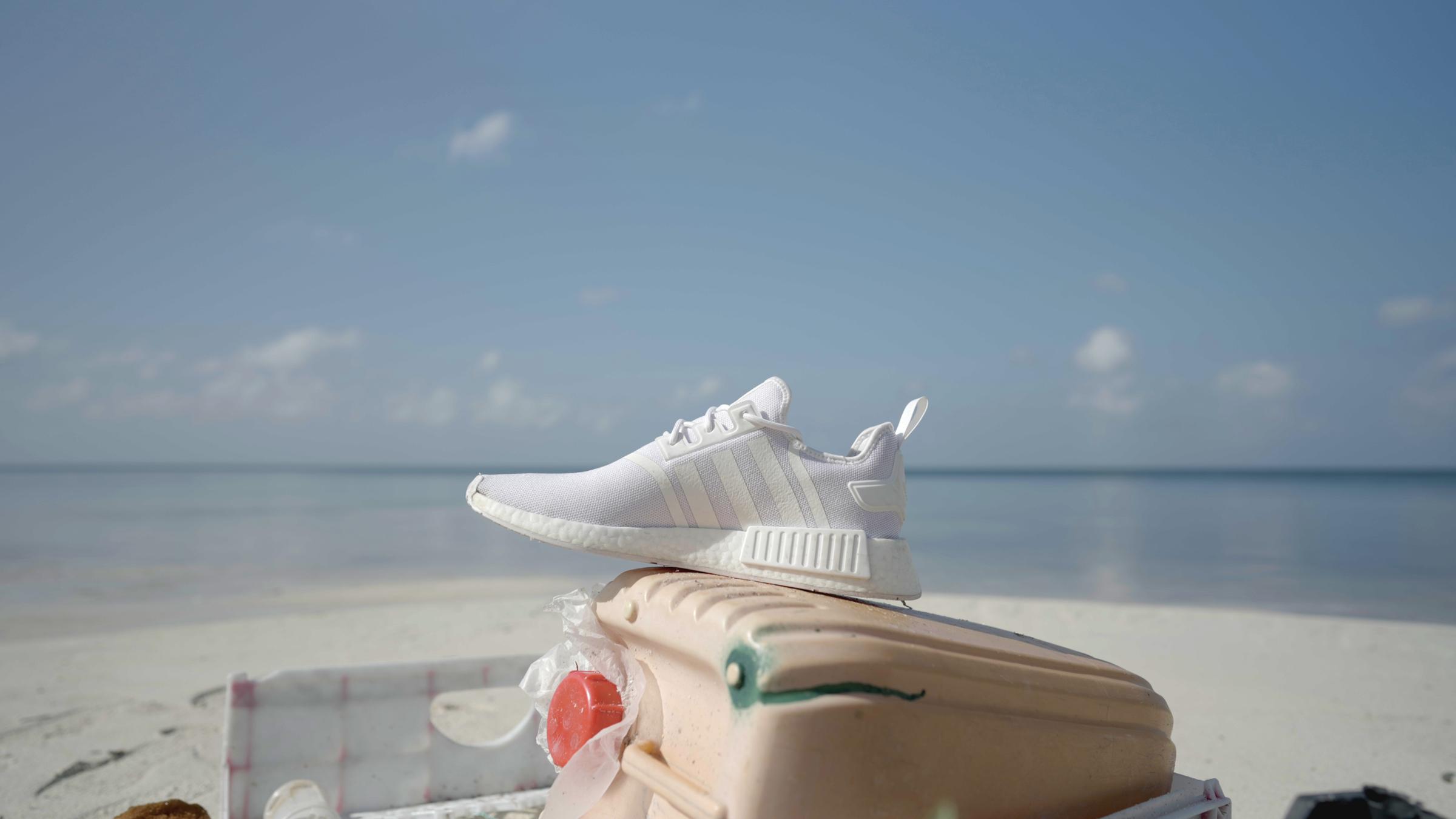 "Greenwashed? Der Schuh aus Meeresplastik": Ein neuer, weißer Sneaker steht auf einem beigefarbenen Kanister am Strand. Im Hintergrund ist das Meer.