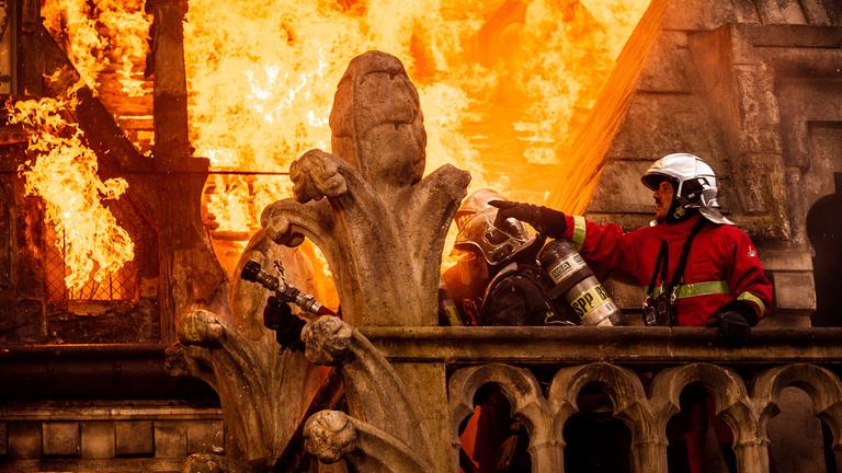 "Notre-Dame in Flammen": Feuerwehr-Chef Jordan (Jules Sadoughi) steht auf einer Balustrade und zeigt seinem Team wo sie das Feuer löschen sollen. Hinter ihnen steht eines der Dächer der Kathedrale komplett in Flammen.