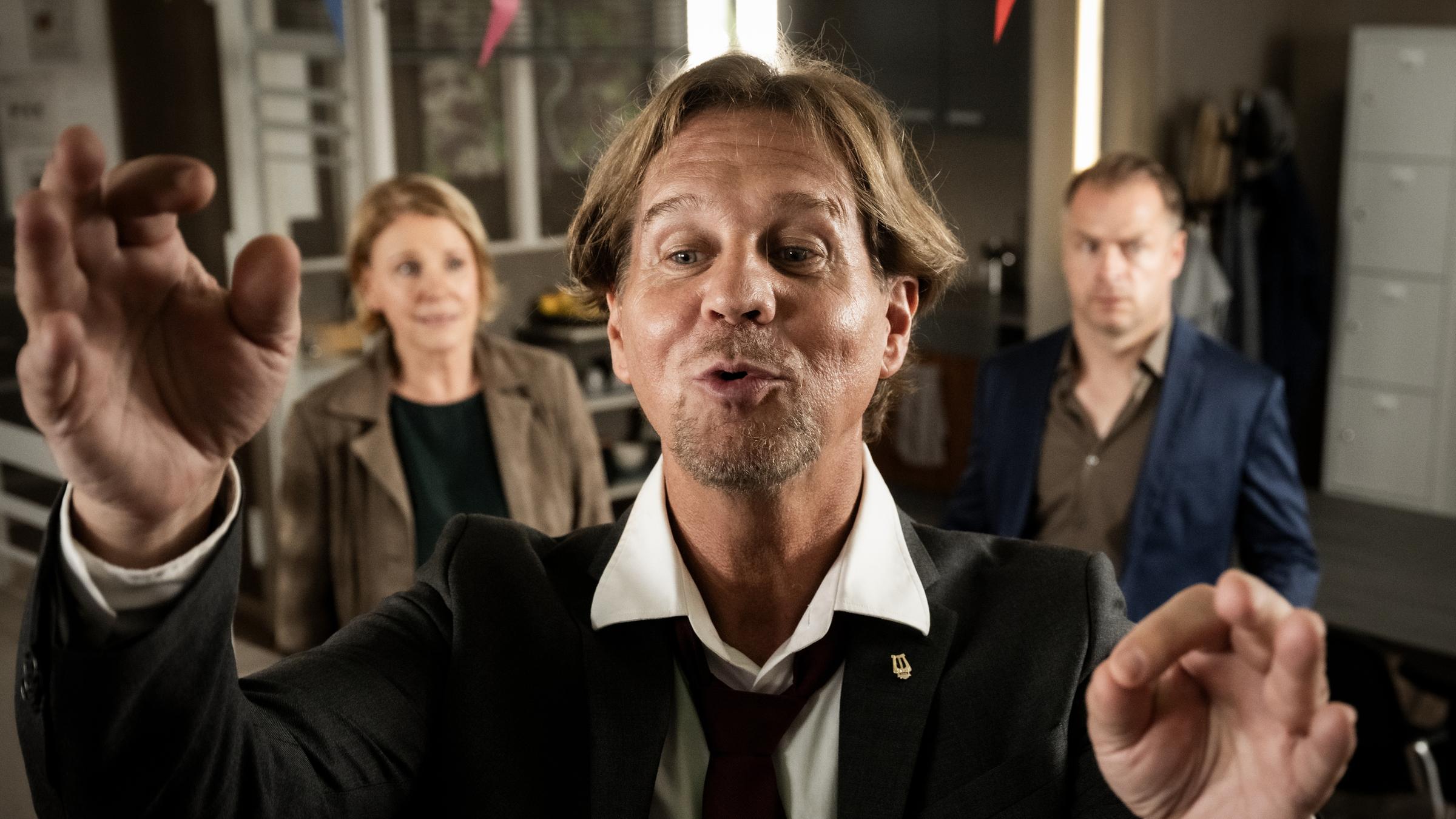 "Marie Brand und die verfolgte Braut": Marie Brand (Mariele Millowitsch) und Jürgen Simmel (Hinnerk Schönemann) stehen im Revier und schauen perplex zur Seite. In der Mitte steht Dr. Engler (Thomas Heinze) mit gespitzten Lippen und dirigiert.