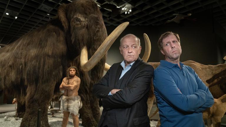 "Wilsberg - Blinde Flecken": Wilsberg (Leonard Lansink) steht Rücken an Rücken mit Ekki (Oliver Korittke) in einem Raum eines Naturkundemuseums. Ihre Arme sind verschränkt und sie werfen sich unfreundliche Blicke zu. Im Hintergrund steht ein großes ausgestopftes Mammut, darunter die Figur eines Neandertalers. Dieser steht ebenso wie Wilsberg und Ekki mit verschränkten Armen da.