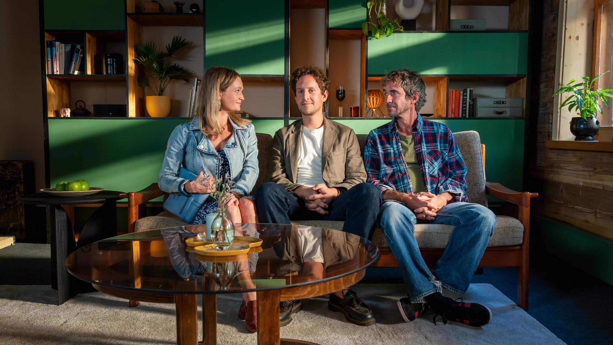 "Familie Anders - Mann Nummer 1": In Fabians (Moritz Treuenfels) Praxisräumen sitzt Fabian Anders (Moritz Treuenfels) zwischen Katja (Natalie Spinell) und Oliver (Felix Hellmann) Koch auf einer Couch. Das Paar schaut Fabian lächelnd an. Im Hintergrund steht eine Schrankwand mit Büchern, Blumen und Dekoobjekten.
