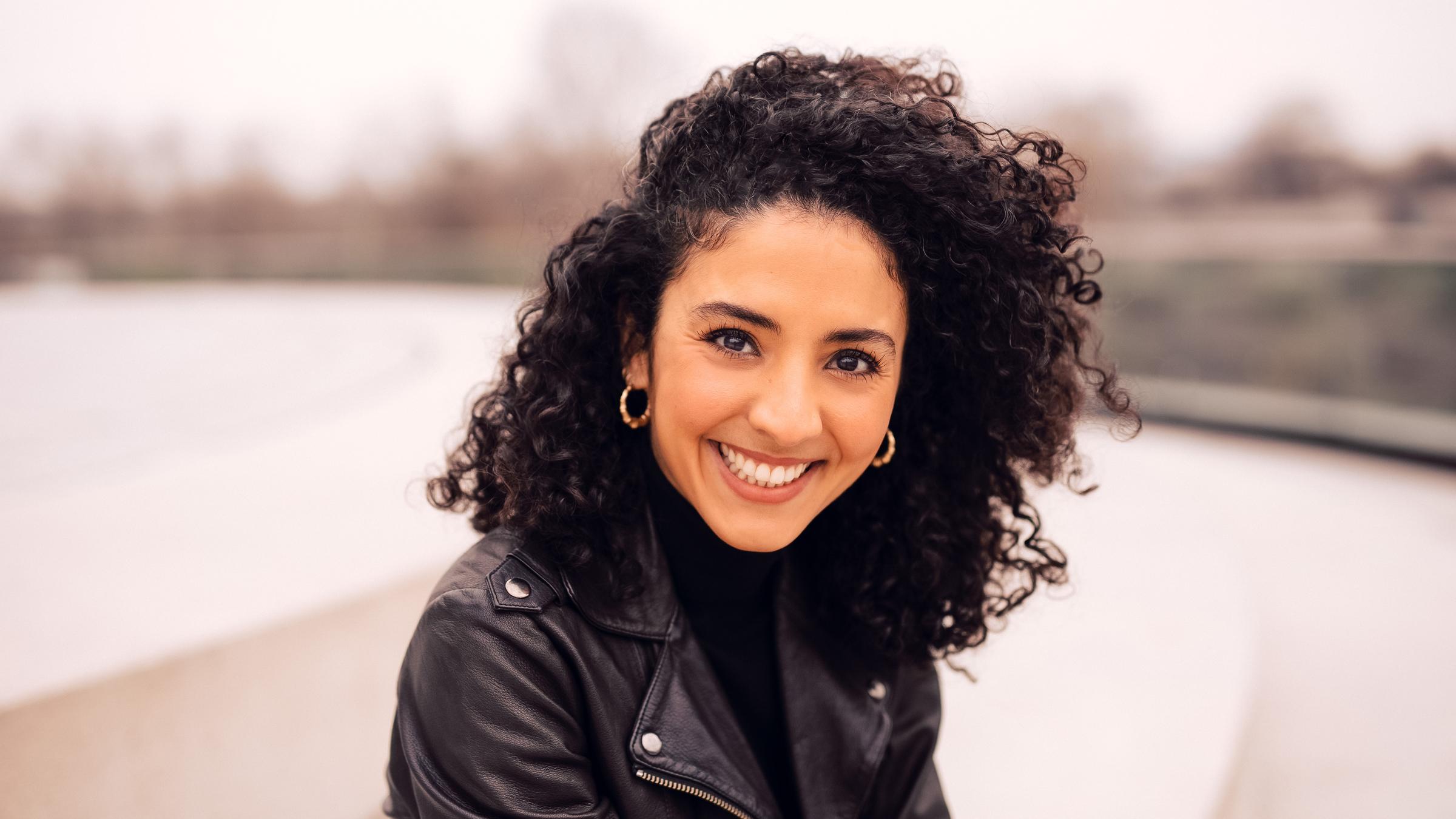 "Verleihung des 60. Grimme-Preises 2024": Auf dem Bild ist eine Frau zu sehen. Sie schaut direkt in die Kamera und lächelt. Die Frau trägt eine schwarze Lederjacke und Ohrringe. Sie ist das Zentrum des Bildes. Der Hintergrund ist unscharf.