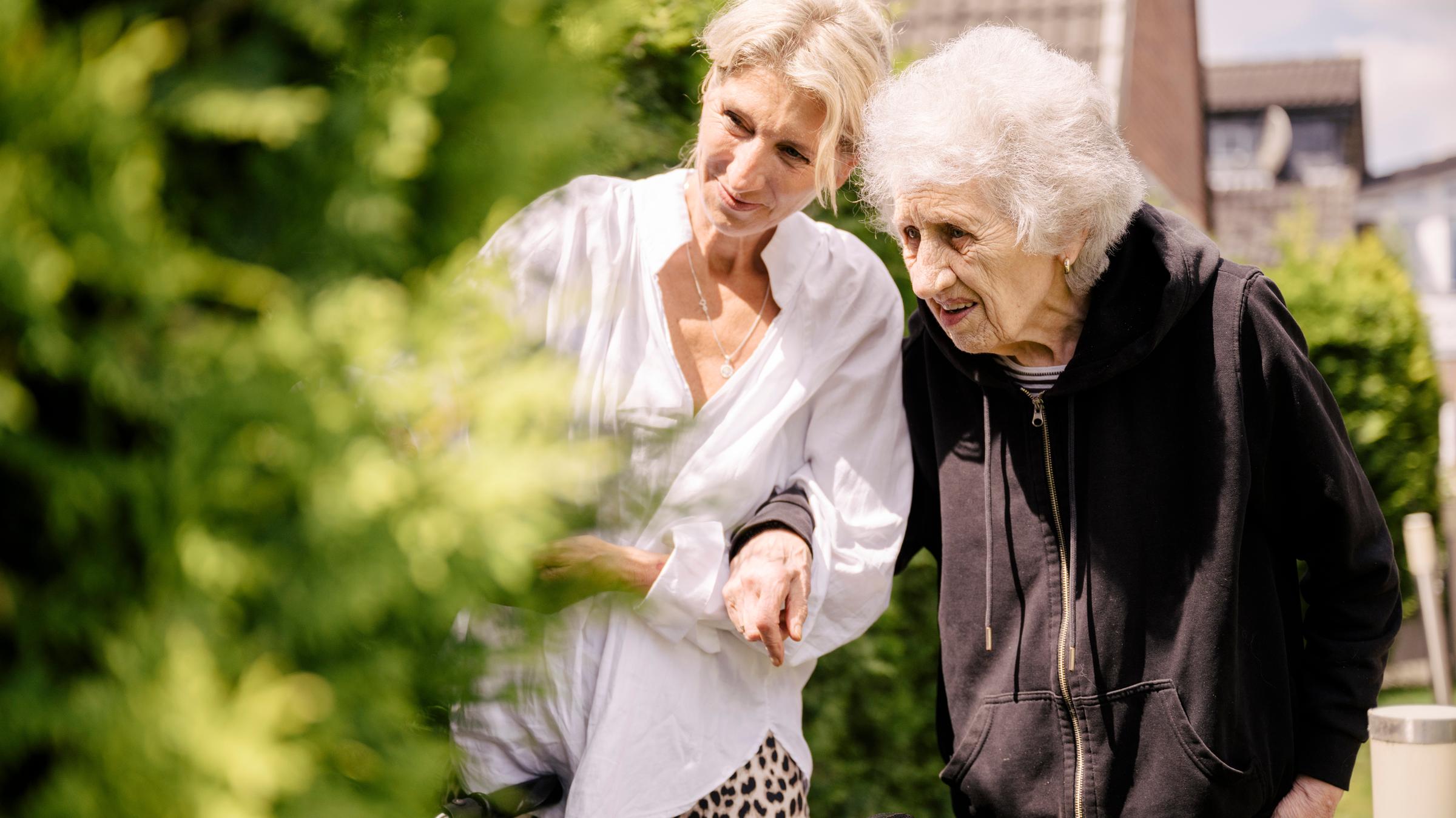 "37°: Ein Zuhause für Vergessende - Betreuung von Demenzkranken": Julia Bernsee, eine Frau im mittleren Alter und ihre Mutter, eine hochbetagte Seniorin, gehen spazieren und betrachten einen grünen Busch.