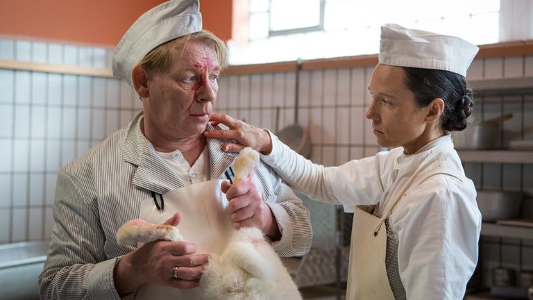 "Karigula": Charly (Ben Becker) und Karla (Sabine Timoteo) stehen nebeneinander. Charly, mit Blutspritzern im Gesicht, hält ein totes weißes Kaninchen, während Karla ihre rechte Hand auf seine Schulter gelegt hat.