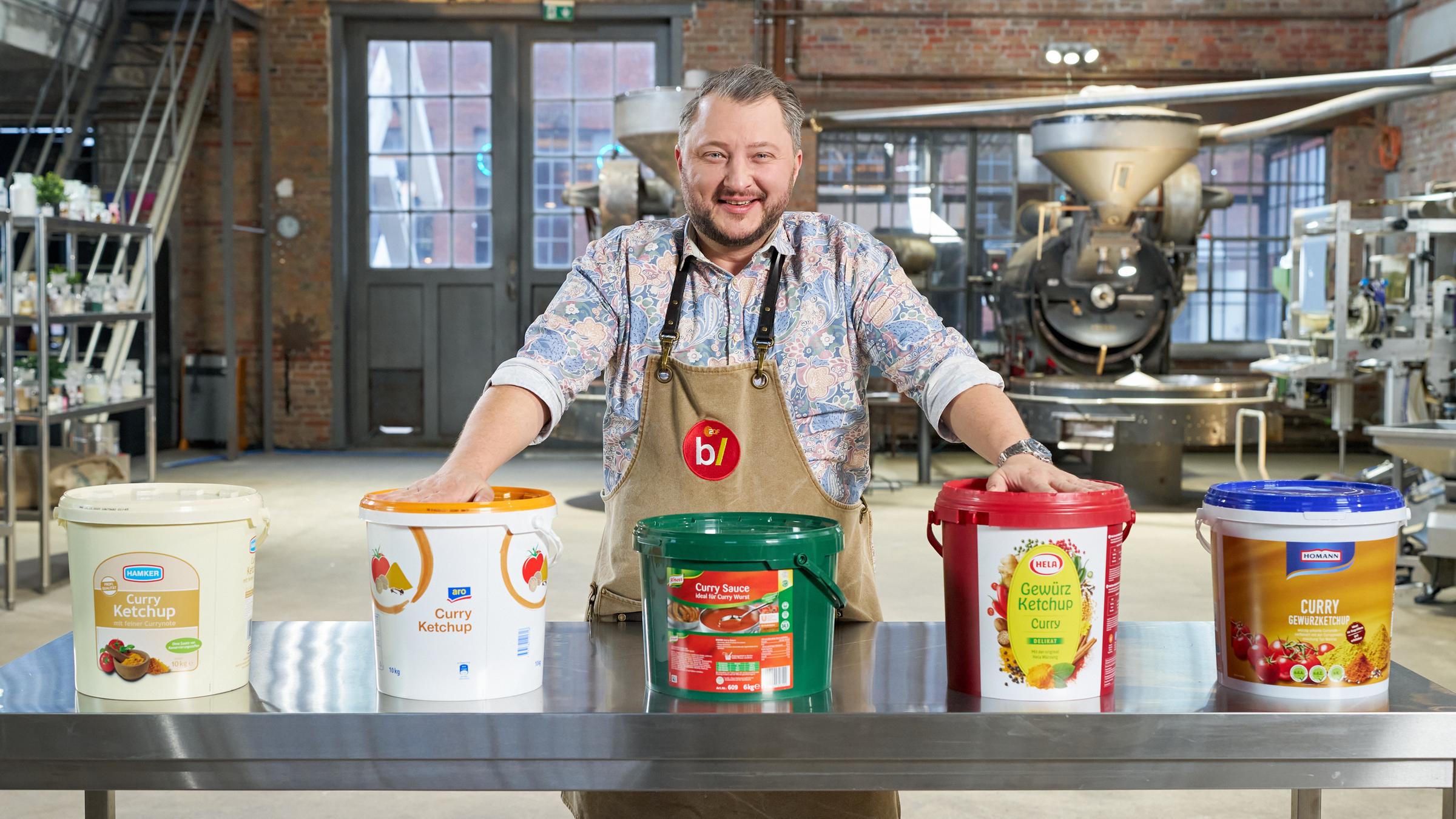 "besseresser: Die Tricks der Gastro-Industrie - Currywurst & Co.": Sebastian Lege steht hinter einem Tisch, auf dem fünf verschiedene XXL-Eimer unterschiedlicher Saucen-Hersteller stehen.
