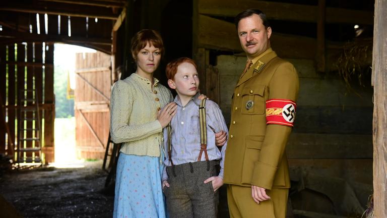 "Das Glaszimmer":  Felix (Xari Wimbauer) steht in einer Scheune zwischen seiner Mutter Anna (Lisa Wagner), die ihre Arme um seinen Hals gelegt hat und Ortsgruppenleiter Feik (Philipp Hochmair), der ihn am rechten Arm feesthält und blickt ihn verunsichert an.