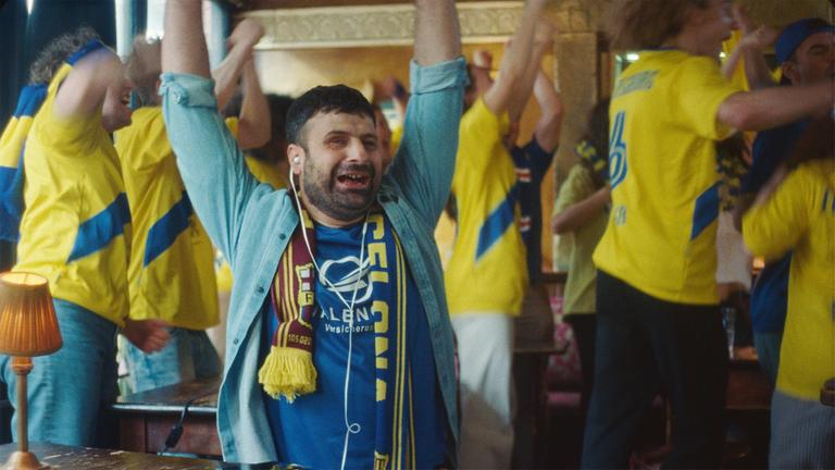 Fans in gelben Trikots strecken die Arme in die Höhe, während sie eine Sportübertragung schauen. Einer von den Fans mit einer Sehbeeinträchtigung steht jubelnd im Vordergrund und trägt In-Ear-Pods.