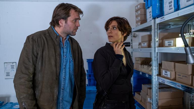 "In Wahrheit - Zwischen Recht und Gerechtigkeit": In einem Lagerraum: Judith Mohn (Christina Hecke) und Alain Montand (Pierre Kiwitt) stehen vor einem Metallregal, in dem sich einige Kartons stapeln. Sie blicken sich in die Augen. Judith hält zudem eine Taschenlampe in ihrer rechten Hand und leuchtet damit in das Regal.