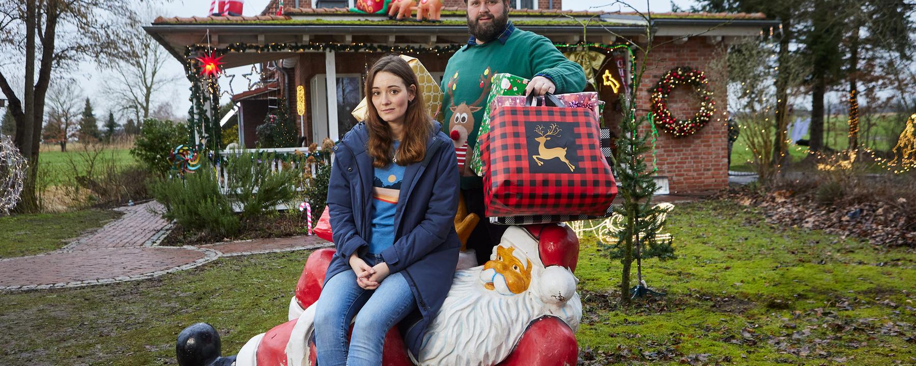 Liane (Maria Ehrich) sitzt auf einer liegenden Weihnachtsmann-Dekoration auf dem Rasen vor einem weihnachtlich geschmückten Haus. Sie schaut amüsiert und blickt in die Kamera. Hinter ihr steht Hinnerk (Antoine Monot), der viele große, bunte Weihachtsgeschenke unter den Armen hält Auch er schaut lachend in die Kamera.