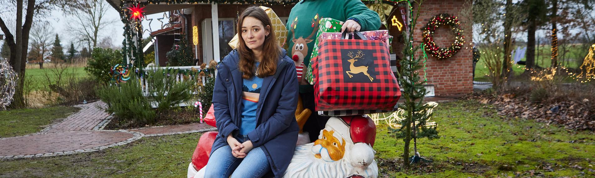 Liane (Maria Ehrich) sitzt auf einer liegenden Weihnachtsmann-Dekoration auf dem Rasen vor einem weihnachtlich geschmückten Haus. Sie schaut amüsiert und blickt in die Kamera. Hinter ihr steht Hinnerk (Antoine Monot), der viele große, bunte Weihachtsgeschenke unter den Armen hält Auch er schaut lachend in die Kamera.