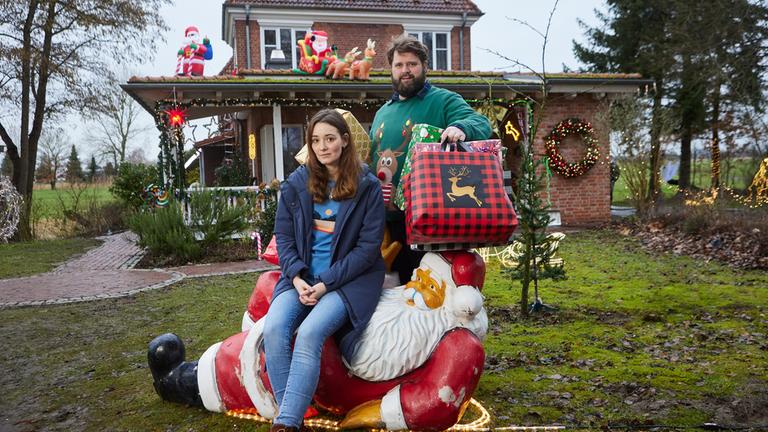 Liane (Maria Ehrich) sitzt auf einer liegenden Weihnachtsmann-Dekoration auf dem Rasen vor einem weihnachtlich geschmückten Haus. Sie schaut amüsiert und blickt in die Kamera. Hinter ihr steht Hinnerk (Antoine Monot), der viele große, bunte Weihachtsgeschenke unter den Armen hält Auch er schaut lachend in die Kamera.