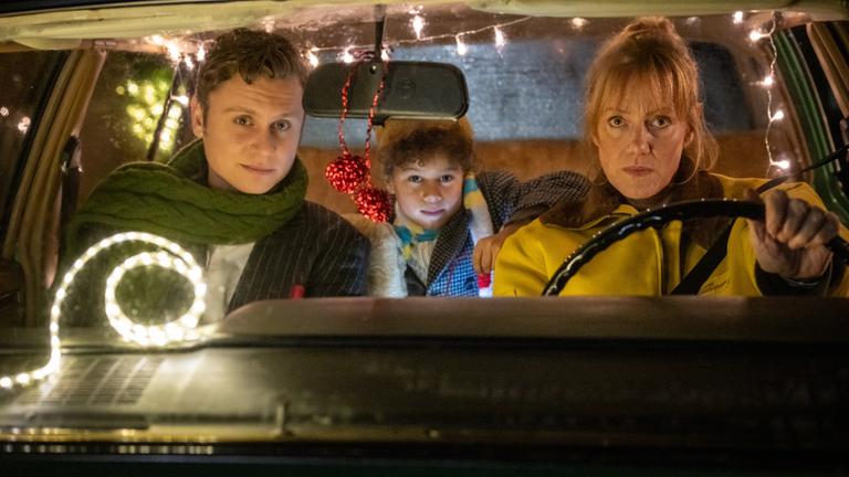 "Nelly und das Weihnachtswunder": Sonny (Rafael Gareisen), Nick (Joah Lion Goetze) und Nelly (Anna Schudt) sitzen in einem Auto, das mit Lichtergirlanden im Inneren geschmückt ist. Nelly sitzt am Steuer, Sonny sitzt neben ihr, Nick sitzt hinter den beiden und hat einen Arm auf Nellys Rückenlehne gelegt. Alle schauen interessiert nach vorne.