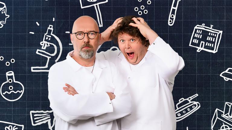 "Der satirische Jahresrückblick 2024": Andreas Wiemers und Werner Doyé stehen, in weißen Kitteln gekleidet, nebeneinander und blicken in die Kamera. Werner Doyé hält die Hände auf dem Kopf und schaut erstaunt.