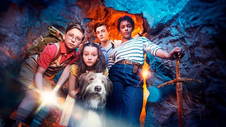 "Fünf Freunde - Der Fluch der Felseninsel": Dick (Kit Rakusen) , Anne (Flora Jacoby Richardson), Julian (Elliott Rose.), George (Diaana Babnicova) und Timmy stehen in einer Höhle und blicken nach unten in die Kamera. Dick leuchtet mit einer Taschenlampe in die Kamera, während George sich mit ihrer linken Hand auf ein Schwert stützt.