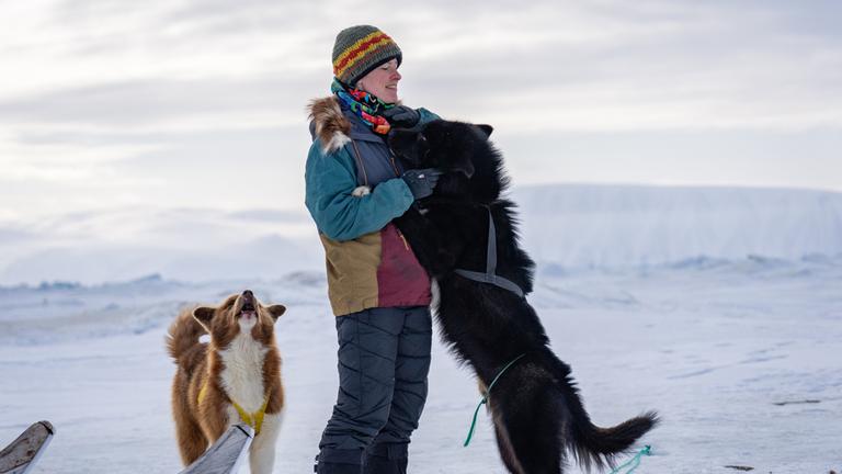 "Grönland": Josefin Kuschela steht in einer Schneelandschaft. Ein schwarzer Schlittenhund steht auf den Hinterläufen und hat seinen Kopf an die Schulter von Josefin gelegt.  Hinter den beiden steht ein weißbrauner, heulender Schlittenhund.