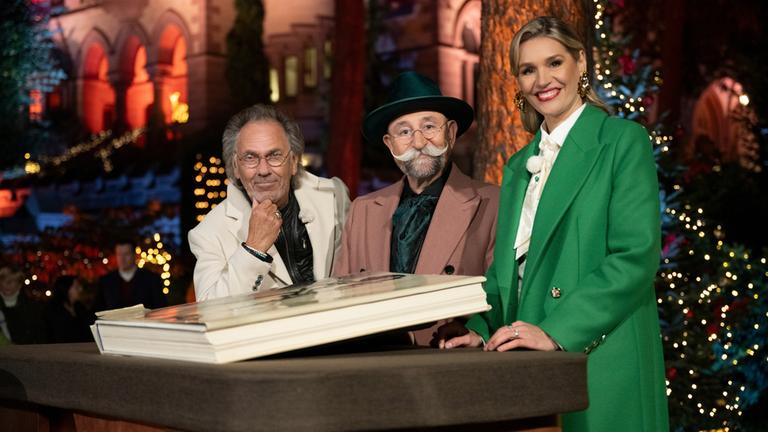 "Bares für Rares - Deutschlands größte Trödelshow": Hugo Egon Balder, Horst Lichter und Dr. Bianca Berding stehen hinter dem Expertisentisch und lächeln in die Kamera. Vor ihnen liegt ein großes Buch. Im Hintergrund ist ein weihnachtlich dekorierter Park und Schloss Drachenburg zu erkennen.