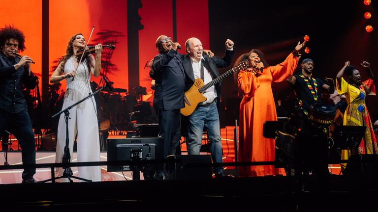 "The World of Hans Zimmer": Hans Zimmer steht mit einer Gitarre zwischen sechs Solistinnen und Solisten auf der Bühne und alle strecken begeistert einen Arm hoch.